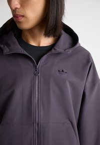 Purple zip-up hooded jacket in smooth fabric. Features an embroidered logo on the left chest and a zip closure with a round pull.