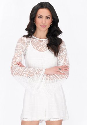 Nainen, jolla on pitkät tummat laineikkaat hiukset, valkoinen pitsinen pitkähihainen paita ja shortsit, seisoo käsivarret ristissä valkoista taustaa vasten.