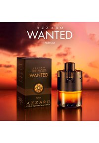 Sort og ravfarvet Azzaro The Most Wanted parfume flaske og æske på reflekterende overflade med en orange solnedgangshimmel i baggrunden.