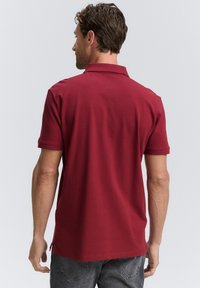 Camisa polo burdeos de tela suave y texturizada; cuenta con un cuello y mangas cortas; costuras visibles a lo largo del dobladillo y las mangas.