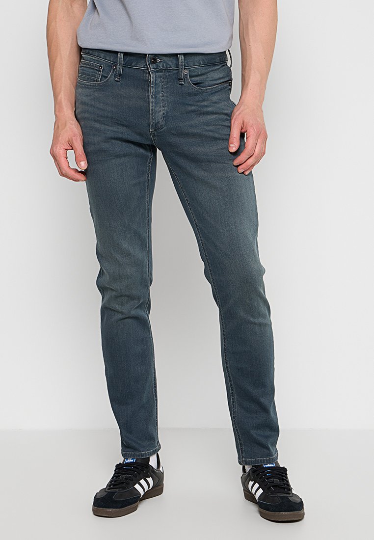 Slim-fit donkere spijkerbroek met een vervaagde wassing, vijf zakken, metalen hardware, riemlussen en een zachte textuur, gecombineerd met zwarte sneakers.