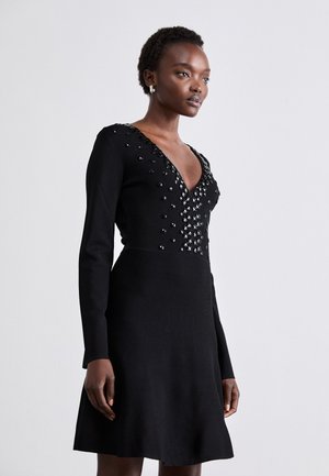 Robe noire à manches longues et décolleté en V profond. Présente des embellissements en perles au niveau du buste et une jupe évasée. Texture lisse et design ajusté.