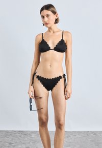 Μαύρο μπικίνι με κυματιστές άκρες, διαθέτει τριγωνικό τοπ και κάτω μέρος με δέσιμο στο πλάι. Το μοντέλο κρατά γυαλιά ηλίου και στέκεται μπροστά σε ανοιχτό φόντο.