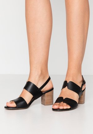 Jambes portant des sandales à talons carrés en cuir noir, à bout ouvert, avec de larges brides et des boucles autour de la cheville, sur un sol clair.
