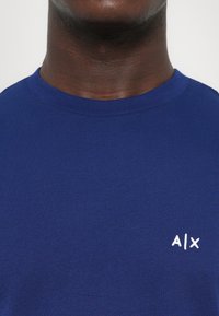 Moški nosi modro majico s krožnim izrezom, na levi strani prsi z majhnim belim logotipom "A|X", nasproti enobarvne bele podlage.