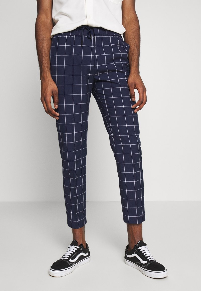 Homme portant un pantalon court à carreaux bleu marine avec une ceinture élastique et des baskets noires à lacets blancs, debout devant un fond uni.