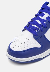 Modro-bílá teniska s hladkým koženým svrškem, perforovanou špičkou, texturovanými akcenty a plochými tkaničkami. Zaoblená špička a kontrastní podrážka.