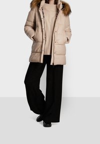 Beige puffjacka med huva i konstfuskpäls, med horisontell sömnad, dragkedja fram och en avslappnad passform, bärs över en krämfärgad tröja och svarta byxor.