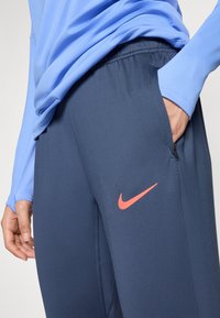 Pantalons de sport Nike en tissu lisse bleu marine ; dotés d'une poche latérale et d'un logo swoosh corail sur la cuisse.