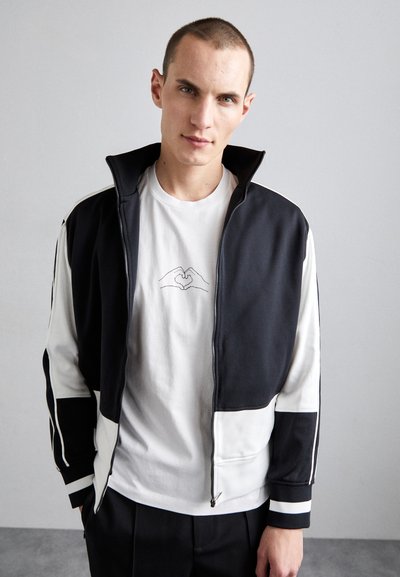 Zip-up jacket in navy and white, with color-block design. Features a high collar, striped accents, and a relaxed fit over a white graphic t-shirt.