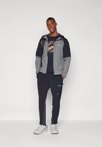 Meeste välivorm, mis sisaldab halli ja tumesinist kapuutsiga jope, tumesiniseid pükse helkurdetailidega ja heledaid tossusid.