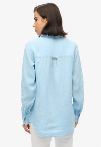 Jasnoniebieska, długopisowa koszula z miękką teksturą, kołnierzykiem i mankietami na guziki. Posiada centralny szew z tyłu oraz akcentującą metkę w okolicy szyi.