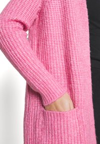 Handen inskjuten i fickan på en lång, ribbad rosa stickad cardigan som bärs över en svart outfit.
