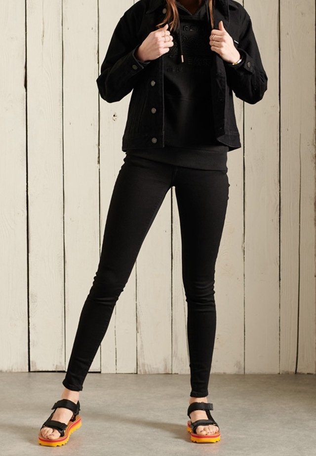 Persoon in een zwarte jas, hoodie en leggings, staand met de benen uit elkaar, in zwarte sandalen met oranje zolen tegen een houten muur.