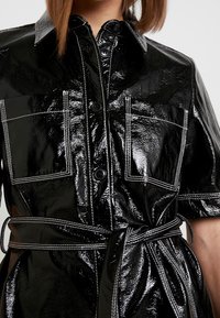 Svart, blanka skjortklänning med korta ärmar, utrustad med stora bröstfickor, vit sömnad och ett knytbälte i midjan. Knappar längs fram.