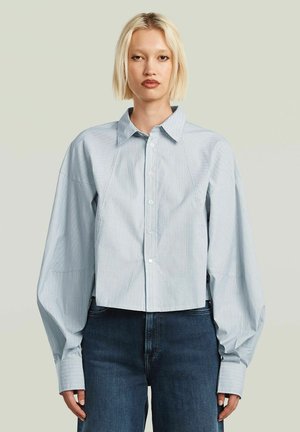 Blondine in een lichtblauw gestreept oversized blouse met knopen en lange mouwen en donkere blauwe spijkerbroek, staand tegen een effen achtergrond.