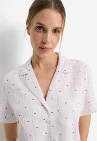 Πυτζάμα με κοντά μανίκια σε λευκό με ροζ ρίγες, διασκορπισμένες μικρές κόκκινες καρδιές, μία τσέπη και γιακά με πέτο. Κατασκευασμένη από μαλακό ύφασμα.