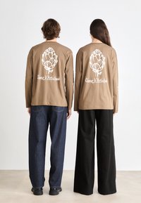 Brown long-sleeve shirts feature a white artichoke graphic on the back. Models wear relaxed-fit jeans, one in dark blue, the other in black.