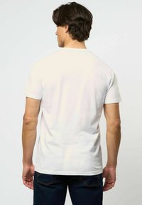 Vīrietis ar tumšiem matiem, tērpies vienkāršā baltā īsroku krekliņā un tumšās džinsās, stāvot ar muguru pret vienkrāsainu gaišu fonu.