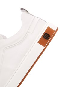 Białe tenisówki z gładką skórzaną cholewką, pętelką na pięcie, białą środkową podeszwą oraz brązową gumową podeszwą z czarnym logo po boku.