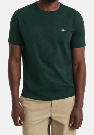 Homme portant un t-shirt vert foncé à manches courtes avec un petit logo sur la poitrine et un pantalon beige, debout devant un fond uni.