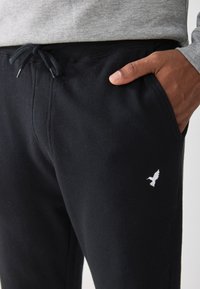 Black sweatpants made of cotton with a drawstring waist, featuring side pockets and a white bird logo on the left thigh.