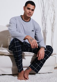 Camisa gris de manga larga con cuello negro y gráfico de ciervo, combinada con pantalones de pijama de cuadros en tono azul marino. Modelo sentado en un sofá claro.