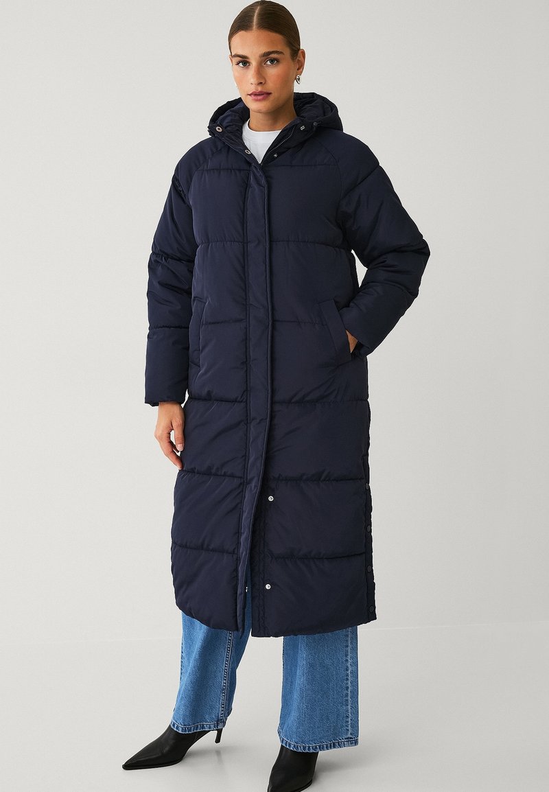 Marineblauwe gewatteerde jas met capuchon, met een lange snit, geborduurde textuur en drukknoopsluiting. Gecombineerd met wijde spijkerbroeken en enkelboots.