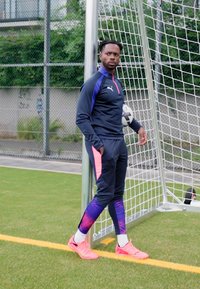 Schwarzes Sportanzug mit lila und pinken Akzenten; beinhaltet ein enganliegendes Oberteil und tapered Hose. Helle pinke Fußballschuhe, einen Fußball haltend.