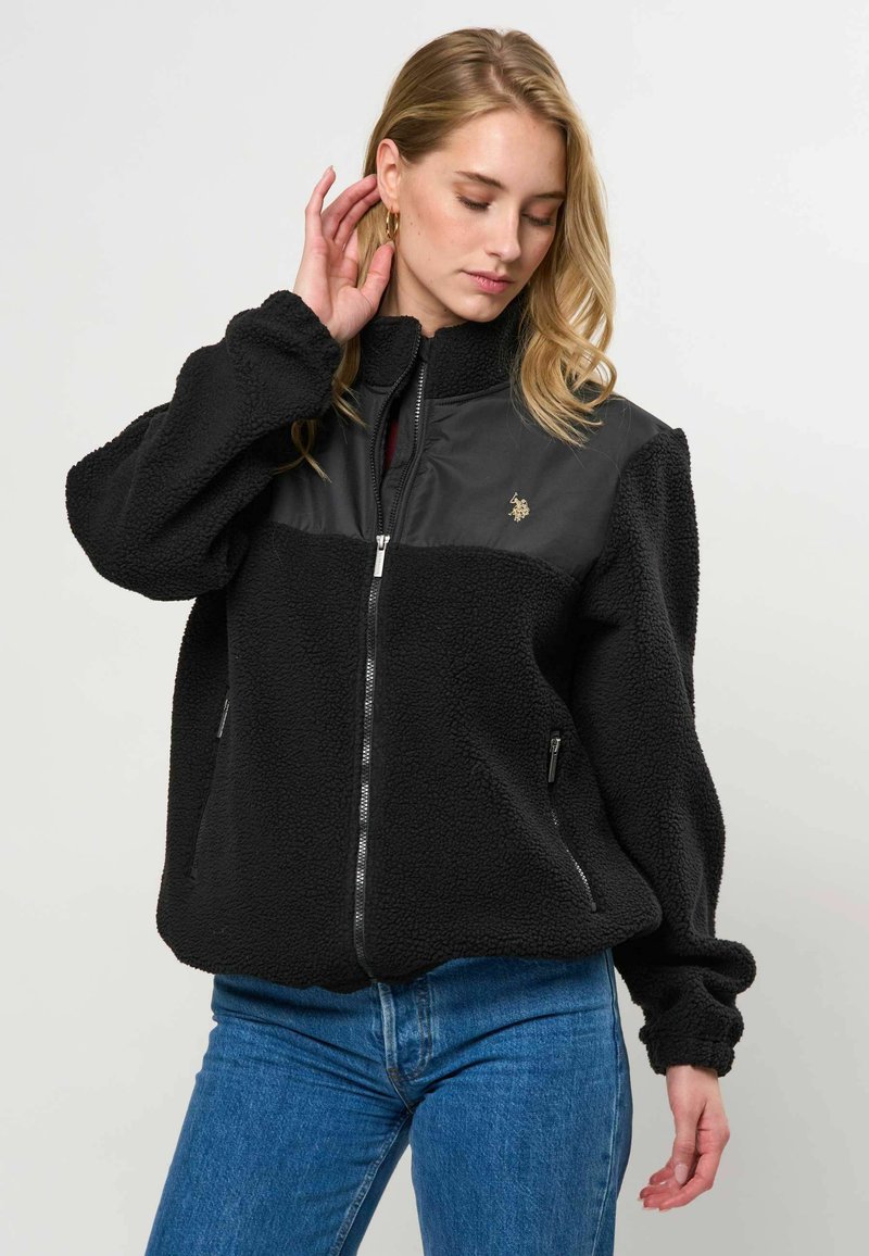 Jeune femme portant une veste polaire noire avec fermeture éclair et empiècement en simili cuir, associée à un jean bleu, posant avec la main près de l’oreille.