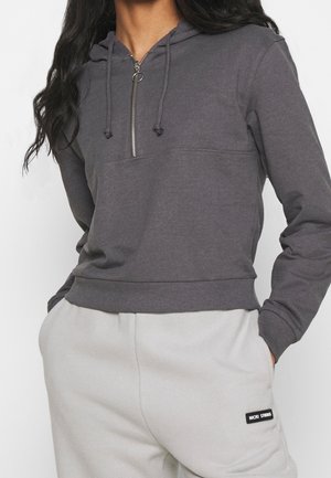 Femme portant un sweat à capuche gris foncé à fermeture éclair partielle et un pantalon de survêtement gris clair, mains dans les poches, cadrée aux épaules et aux hanches.