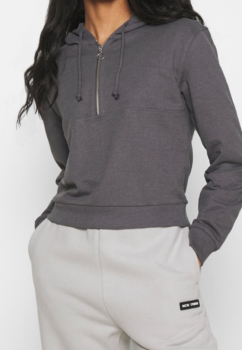 Femme portant un sweat à capuche gris foncé à fermeture éclair partielle et un pantalon de survêtement gris clair, mains dans les poches, cadrée aux épaules et aux hanches.