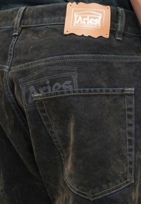 Jeans din catifea neagră cu cusături contrastante, având un patch din piele al brandului Aries pe spate și un logo subtil pe buzunar.