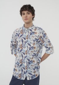Camicia a maniche lunghe con bottoni, caratterizzata da un motivo floreale blu e marrone su un tessuto leggero, con maniche arrotolate e colletto alla mandarino.