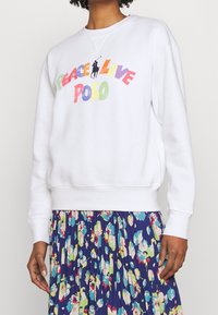 Bílá mikina s nápisem "PEACE LOVE POLO" v barevných písmenech. Má kulatý výstřih a žebrované manžety, nošena přes květinovou plisovanou sukni.