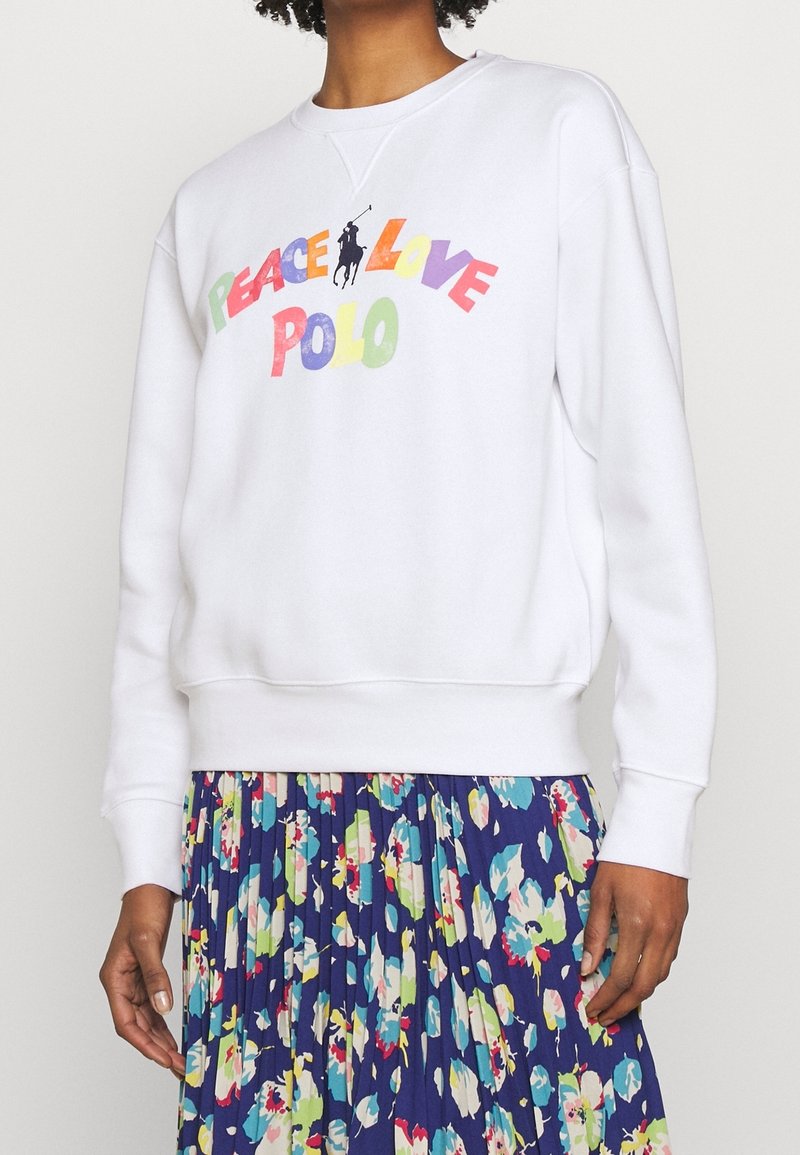 Bílá mikina s nápisem "PEACE LOVE POLO" v barevných písmenech. Má kulatý výstřih a žebrované manžety, nošena přes květinovou plisovanou sukni.