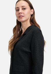 Svart cardigan med texturerad yta, med klassisk krage, framfickor och knappstängning. Slät tyg med en subtil glans.