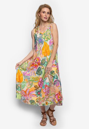 Vrouw die een mouwloze, kleurrijke bloemen maxi-jurk draagt met oranje, roze, groene bladeren en bloemen, poserend in bruine sandalen met bandjes.