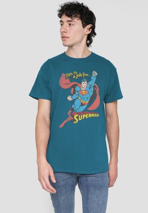 Young man wearing a teal Superman T-shirt with vintage print reading "This is a job for Superman," looking to his left against a plain background.