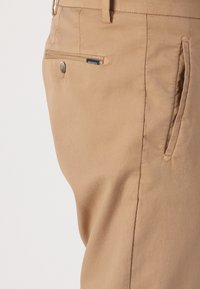 Beige fabric trousers featuring a side pocket with brass button closure and stitched detailing for durability and refined finish.