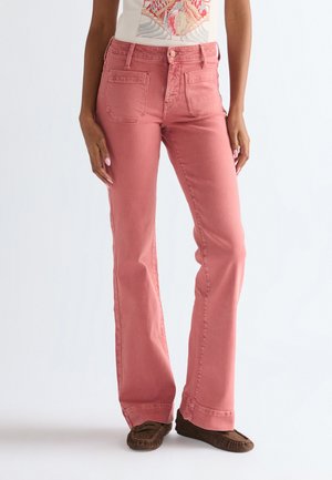 Kobieta ubrana w różowe jeansy z wysokim stanem i prostymi nogawkami z przednimi naszywanymi kieszeniami oraz brązowe mokasyny, stojąca na jednolitym tle.