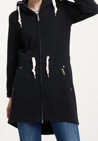 Sweat à capuche long noir à fermeture éclair avec cordons blancs et poches, porté par une personne en jean bleu, bras levé hors du cadre.