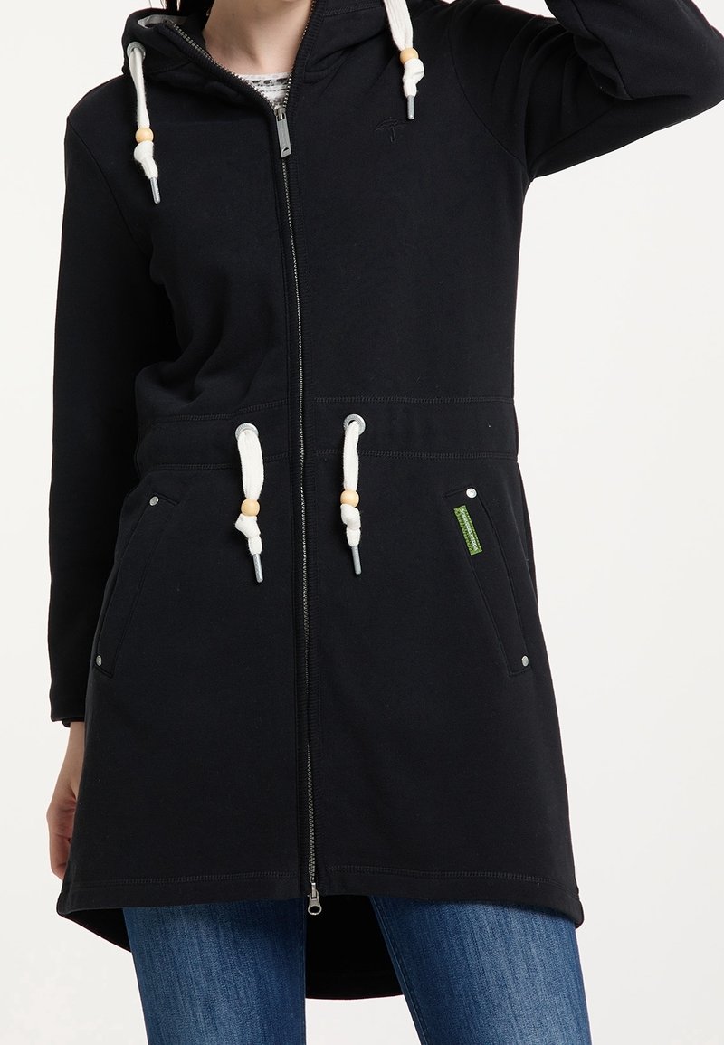 Sweat à capuche long noir à fermeture éclair avec cordons blancs et poches, porté par une personne en jean bleu, bras levé hors du cadre.