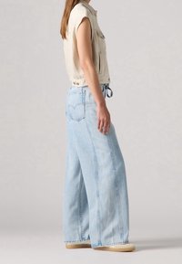 Ljust beige ärmlös topp med framfickor, matchad med vida ljusblå jeans med subtil vertikal sömnad och bakfickor.