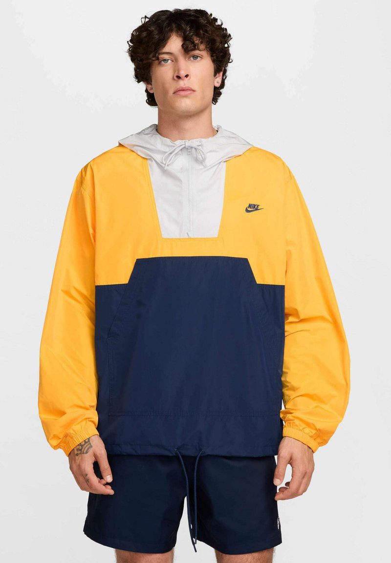 Rompevientos de Nike con parte superior amarilla, capucha gris y sección inferior en azul marino. Incluye un cierre de media cremallera y cordón ajustable en el dobladillo.