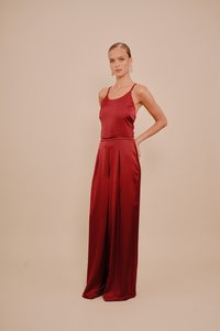 Kvinne som står i en ermeløs burgunder sateng jumpsuit med vide ben foran en enkel beige bakgrunn.