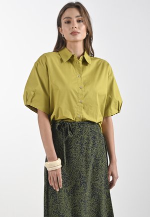 Korte mouwen, kraagshirt in olijfgroen katoen met een knopenfront. Gecombineerd met een donkergepatrooneerde rok en romige armbandjes.