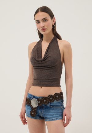 Mujer que lleva un top marrón sin mangas, pendientes grandes de aro, shorts vaqueros azules y un cinturón ancho de cuero con tachuelas y una hebilla grande y redonda.