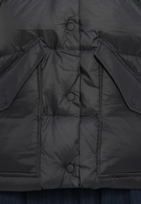 Sort pufferjakke med quiltet design, der har trykknapper og to sidelommer med klapper, lavet af skinnende, glat materiale.