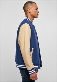 Jachetă varsity albastră cu mâneci bej, guler și manșete ribbate, nasturi tip snap și buzunare laterale, având o textură netedă și un design sportiv.